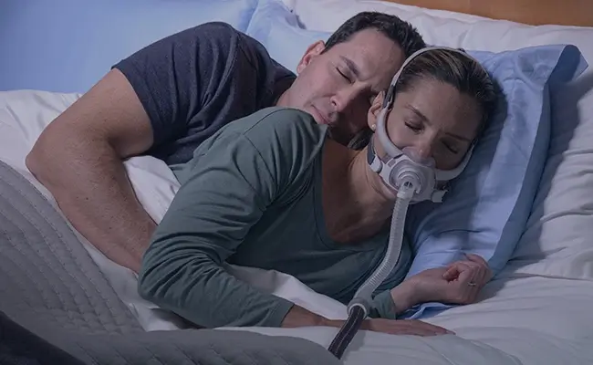 Pariskunta nukkuu lähekkäin vuoteessa, nainen käyttää AirFit F40 CPAP-maskia.