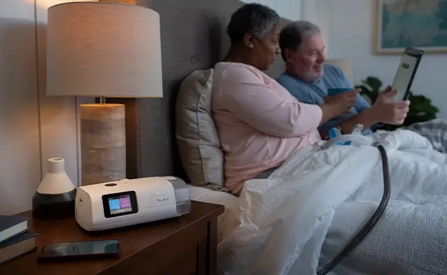 Pariskunta katsoo vuoteessa tablettia. Naisen sylissä on AirCurve 11 ASV PaceWave laitteen maski, joka on kiinnitetty yöpöydällä olevaan laitteeseen.